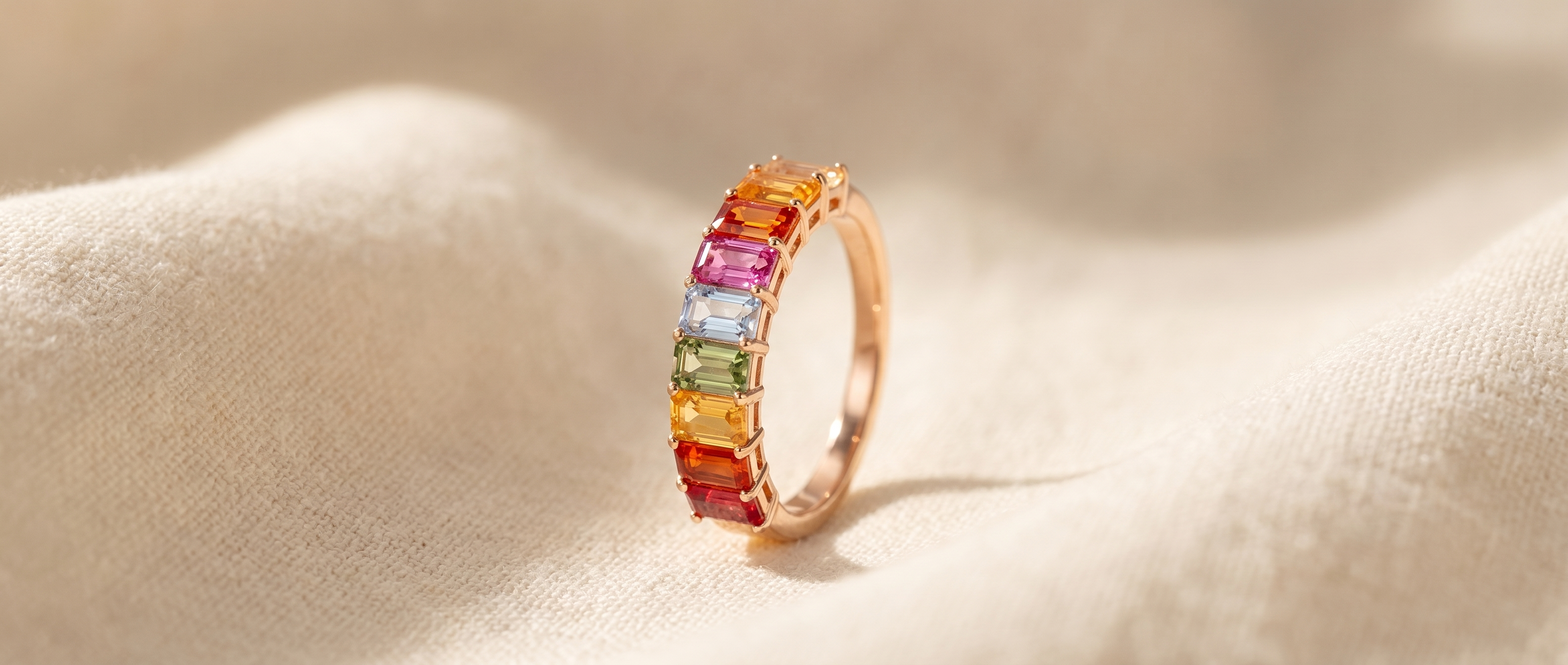 Multi-colour rainbow sapphire half-set ring — stones across the front, plain rose-gold band — at a three-quarter angle on warm cream linen, the Colour collection