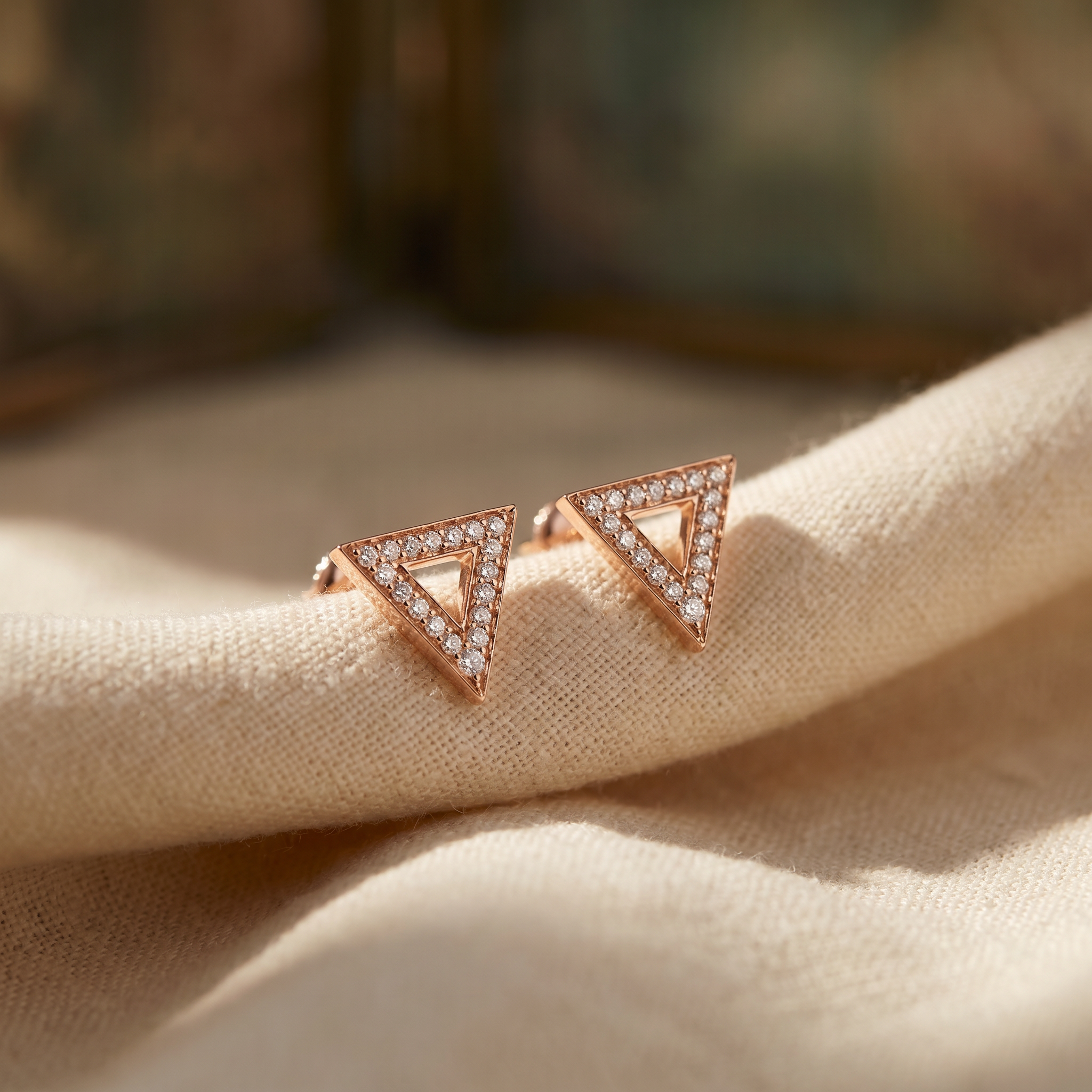 Triangle diamond stud earrings in 18K rose gold, editorial scene on warm linen